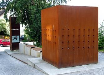 Großer Würfel aus angerostetem Metall mit Lochreihen, als Teil einer Anlage mit Blockelementen am anderen Ende eine Stele mit Informationen