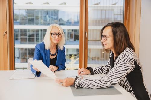 Zwei Mitarbeiterinnen sitzen an einem Bürotisch in modernem Bürogebäude und besprechen eine Unterlage