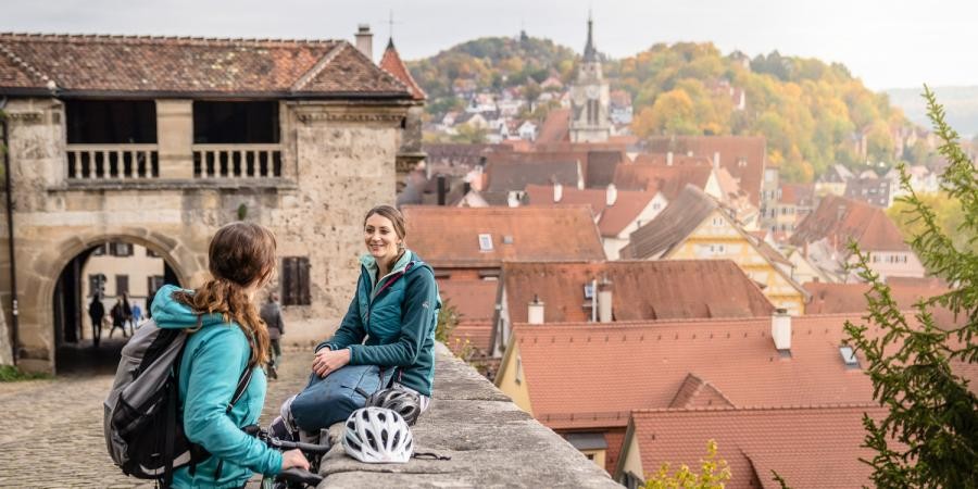 Auf einer Mauer mit Blick über die historische Altstadt von Tübingen sind zwei Frauen im Gespräch, eine Fahrradhelm liegt da auf der Mauer, das Tor des Schlosses ist zu sehen.