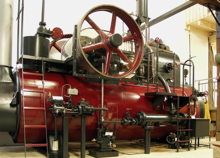 Große Dampfmaschine in einem Innenraum, Dampfkessel mit großem Schwungrad und außenliegenden mechanischen Einrichtungen für die Kraftübertragung