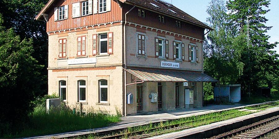 Dreistöckiges Bahnhofsgebäude mit Satteldach, am Bahngleis, der Bahnsteig ist überdacht, das Dachgeschoss mit Holz verkleidet, ein Bahnhofsschild trägt die Beschriftung "Bieringen"