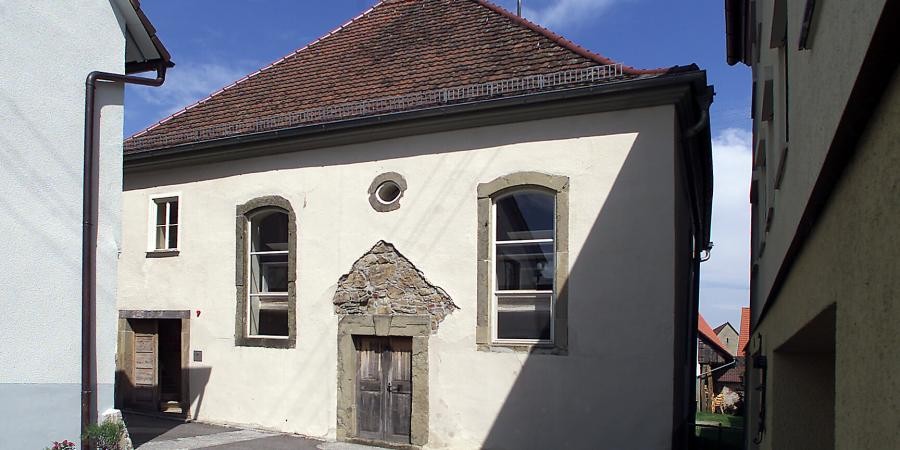 Außenansicht Gedenkstätte Synagoge Baisingen: kleineres walmdachgedecktes einstöckiges Gebäude, Spuren der Beschädigung wie fehlender Putz über der Tür sind zu sehen