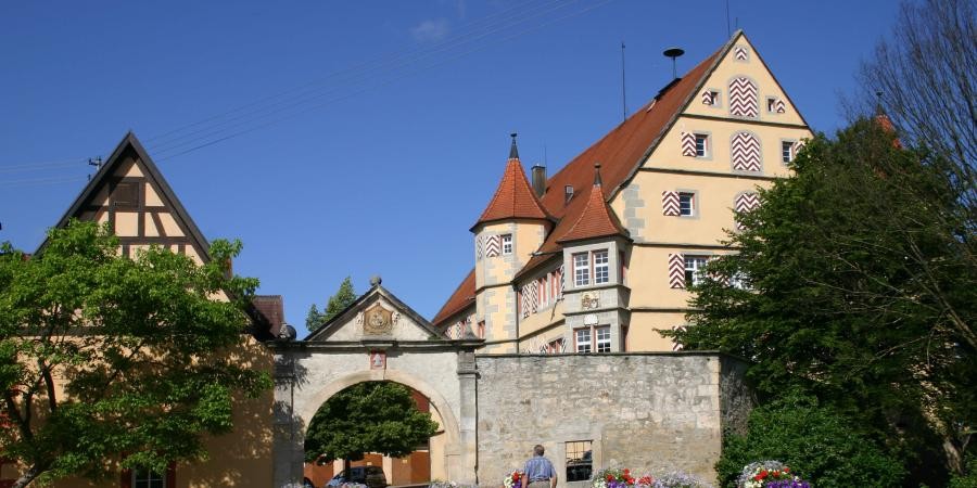 Großes Gebäude mit Satteldach und mehreren Türmen, Zugang durch ein Bogentor in der umgebenden Mauer, nebenstehend ein Gebäude mit Satteldach und Fachwerkgiebel