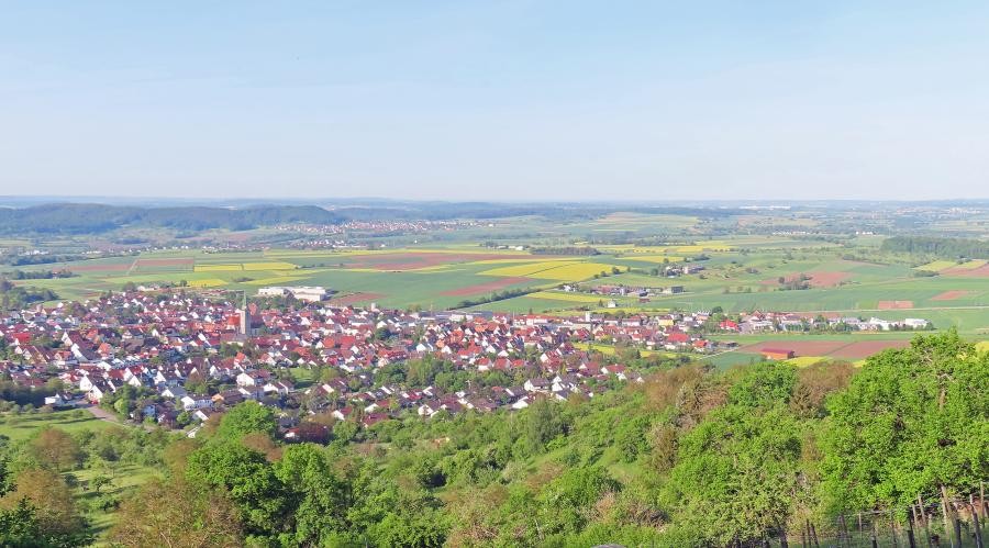 Luftbild einer großen Ortschaft, im Vordergrund bewaldeter Hang, im Hintergrund Kulturlandschaft und die Berge des Schwarzwalds.