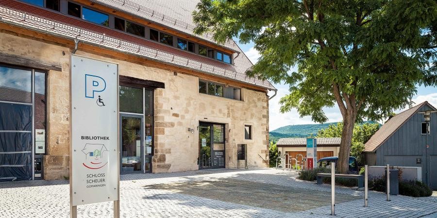 Gomaringen, Bibliothek in der ehemaligen Schlossscheuer Teil eines alten Gebäudes, mit tiefgezogenem Satteldach und Dachgaube, modernisiert teils mit hohen Glasfenstern und Glastüren. Davor gepflasterter Platz mit  Schild "Bibliothek"
