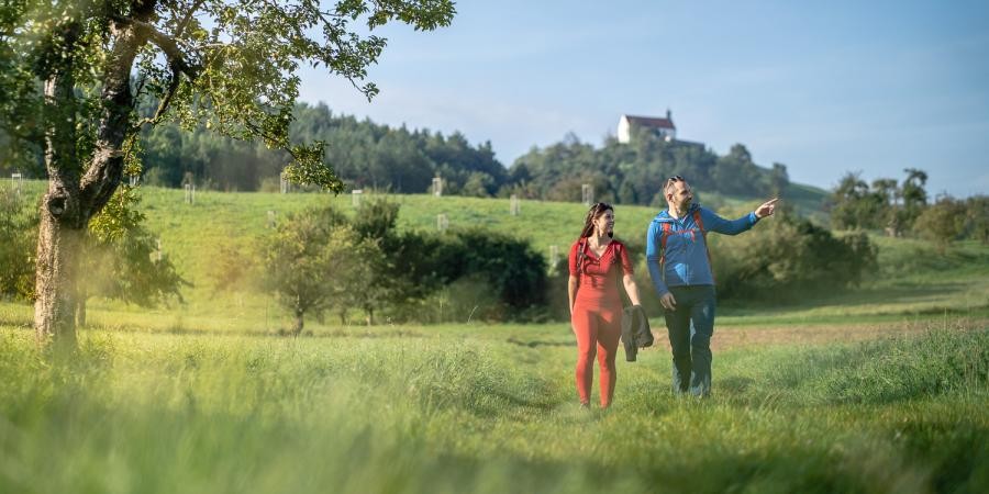Zwei Personen spazieren in einer Wiesenlandschaft, im Hintergrund ein Hügel mit Kapelle