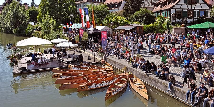 Flussufer mit schwimmender Holzplattform und Schirmen, Holzboote, am  Platz am Ufer viele Menschen, ein Fachwerkhaus der Altstadt