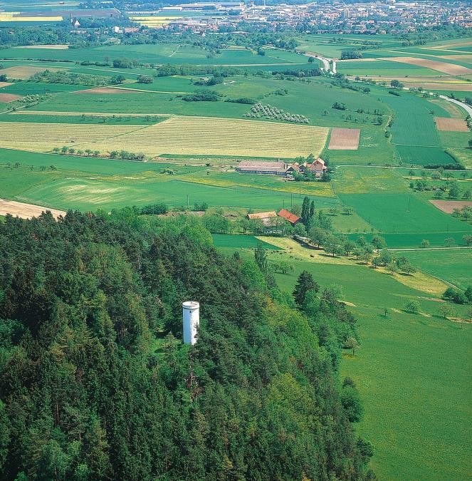 Runder Turm, er ragt aus einem Wald auf einem Hügel, umgebend Wiesen und Felder mit Gehöften, im Hintergrund eine Stadt