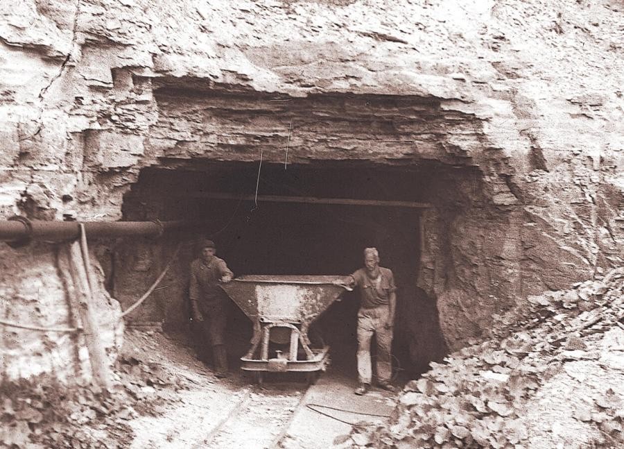 Eine Tunnelöffnung in einem Gesteinshang, zwei Arbeiter schieben eine auf Schienen laufende Lore