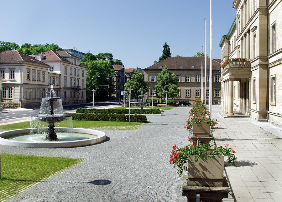 Teil eines großen Gebäudes und säulengestütztem vorkragendem Eingang, großer Vorplatz mit Brunnen, umgebend Straßen und mehrere große Häuser