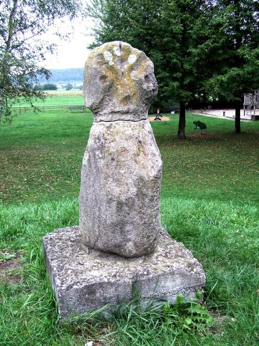 Teil eines grasbewachsenen Hügels, darauf eine Steinfigur auf einem Sockel, mit Körper und Kopf mit Vertiefungen wie Augen und Mund und Einkerbunden
