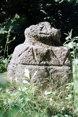 Replik einer figürlichen Steinstele, am Rumpf ist ein großes Zackenmuster umlaufend zu erkennen, am Kopf angedeutete Gesichtsmerkmale