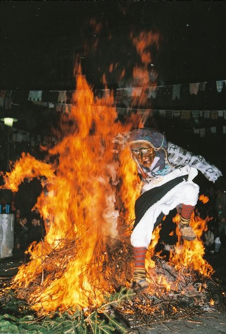 Fasnetsszene: brennender Reisighaufen, ein als Hexe verkleideter Mensch springt bei Dunkelheit durch das Feuer, zuschauende Kinder, über den Platz gespannte Schnüre mit Fasnetslumpen a