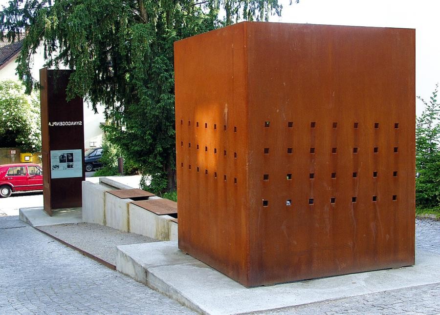 Anlage mit einem Kubus aus Metall mit regelmäßig angeordneten kleineren Öffnungen und rostiger Oberfläche, eine Stele mit Texten und verbindenden  Steinkuben