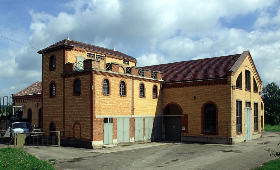 Backsteingebäude mit zwei rechtwinklig aneinandergebauten Gebäudeteilen, Satteldächer, rundbogige Fenster und im Erdgeschoss Tore und