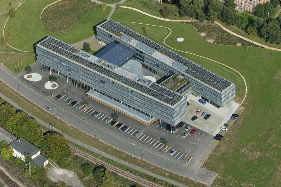 Luftbild: langgestreckte, mehrstöckige Gebäuderiegel mit Querverbindungen, auf den Dächern Photovoltaikpaneele, umgebende Grünflächen 