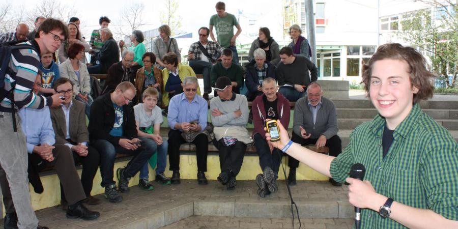 Eine Gruppe von Teilnehmenden sitzt auf Treppenstufen im Freien, eine junge Frau mit Mikrofon zeigt ein elektronisches Gerät