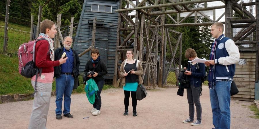 Eine Gruppe von Personen steht vor einem Tor aus einer Holzkonstruktion und einem hölzernen Wachtturm