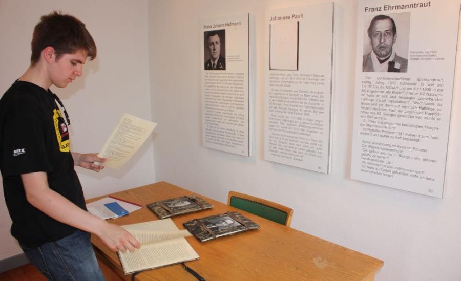 Junger Mann in einem Raum mit Ausstellungstafeln, er blättert an einem Tisch stehend in einem Buch und hält ein Schriftstück in der Hand