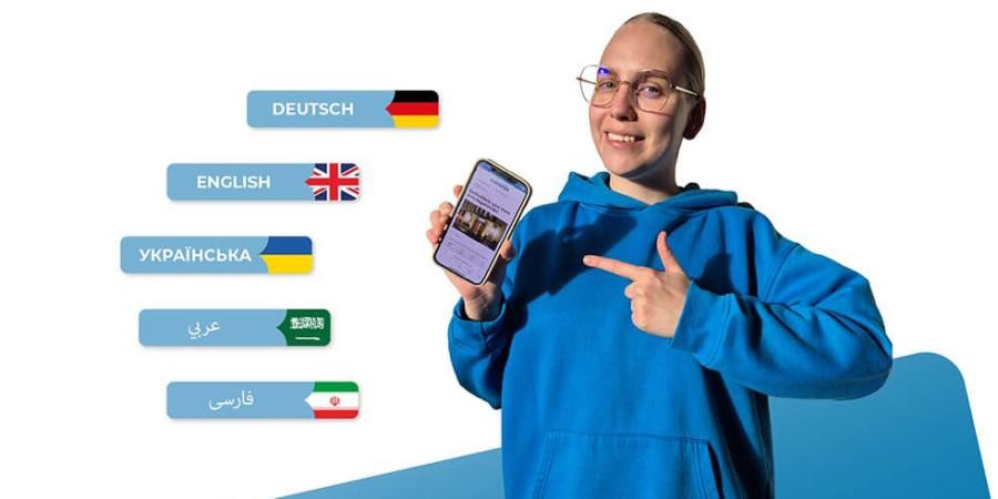 Eine junge Frau zeigt auf ein Handy, auf dem Bildschirm ist die tuenews-App geöffnet, dabei stehen die fünf Sprachen Deutsch, Englisch, Ukrainisch, Arabisch, Dari mit Flaggensymbolen