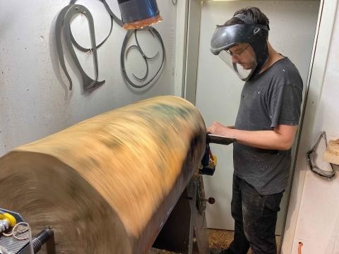 männliche Person mit Gesichtsschutz, die an einem großen Stück eines Holzstammes an einer Drechselbank arbeitet. An der Wand hängt Werkzeug. 