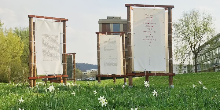 Die bedruckten Planen für die Ausstellung "Wortwiese" sind an dreiseitigen Holzgestellen verschnürt, auf der Wiese beim Landratsamt blühen Narzissen, die Gebäude sind zum Teil zu sehen