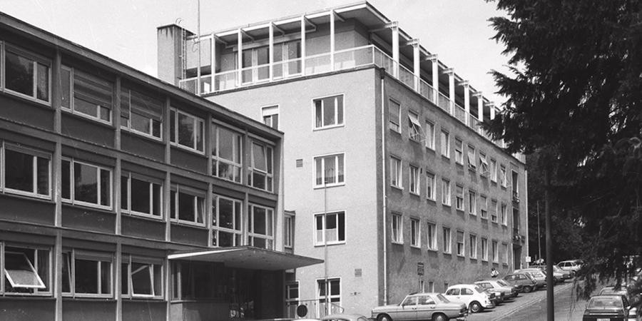 Zwei mehrgeschossige Gebäude mit Flachdach, nebeneinander mit der Längsseite zur Straße stehend, davor geparkte Autos der frühen 1980er Jahre
