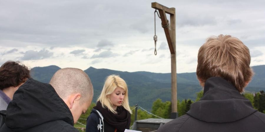 Link zur Rubrik Jugendguides: eine Gruppe von jungen Menschen steht an einem Bergplateau, auf dem Gelände des KZ Natzweiler-Struthof im Elsass, ein Galgen aus Holz steht dort, eine Frau hält ein Manuskript und trägt vor, in der Ferne Berge