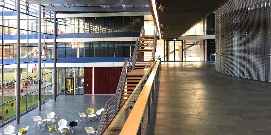 Blick in eine große gläserne Halle von einem Übergang im zweiten Stock aus, in der Halle sind Sitzgruppen, eine Treppe verbindet die Stockwerke