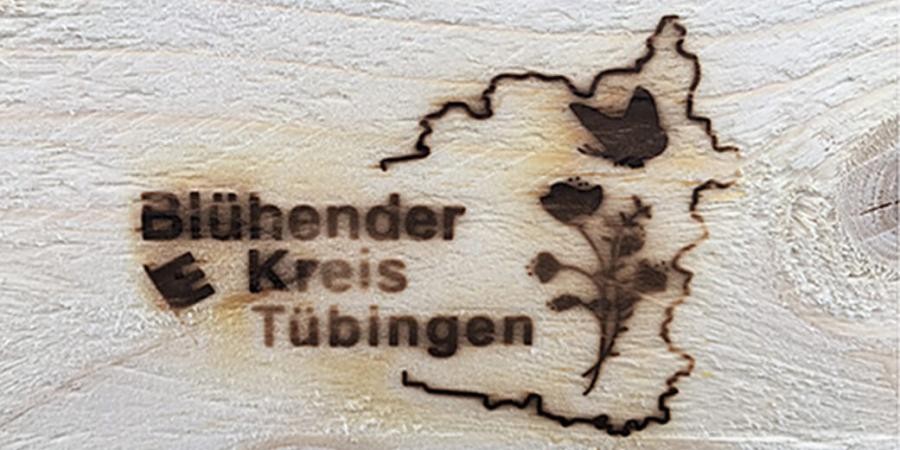 Holz mit eingebranntem Logo: Beschriftung Blühender Kreis Tübingen und Umriss des Landreises mit Blume und Schmetterling