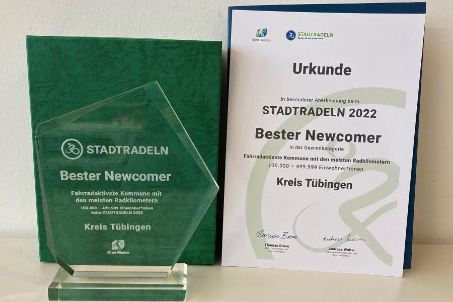 Pokal in Form eines unregelmäßigen Fünfecks aus transparentem Material, daneben eine Urkunde, jeweils mit Beschriftung "Stadtradeln, Bester Newcomer,  Kreis Tübngen