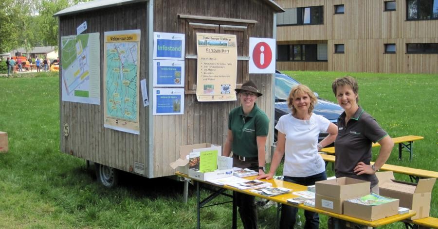 Ein hölzerner Waldarbeiterwagen auf Rädern, mit Plakaten, drei Personen an einem Infotisch bei einer Veranstaltung "Waldtage"
