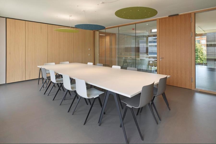 Innenraum mit großem Tisch und ringsum stehenden Stühlen, Gestaltungselemente Holz und Glasflächen