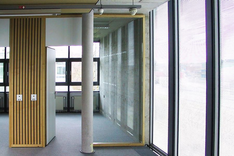 Blick vom Flur in Innenräume, große Glasflächen sind zum Flur und nach außen verwendet, eine Betonsäule und Elemente aus Holz sind zu sehen