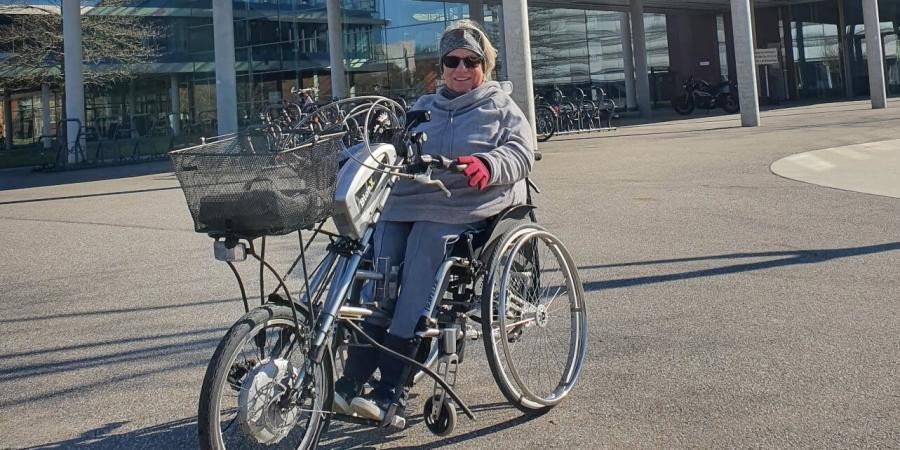 	  Foto von Frau Pflumm. Frau Pflumm ist vor dem Landratsamt. Sie sitzt in einem Fahrzeug. Es ist eine Mischung aus Rollstuhl und Fahrrad.