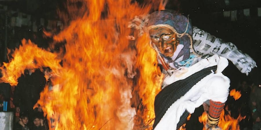 Fasnetsszene: ein als Hexe verkleideter Mensch springt bei Dunkelheit durch ein großes Feuer, im Hintergrund Zuschauende und Schnüre mit Fasnetslumpen