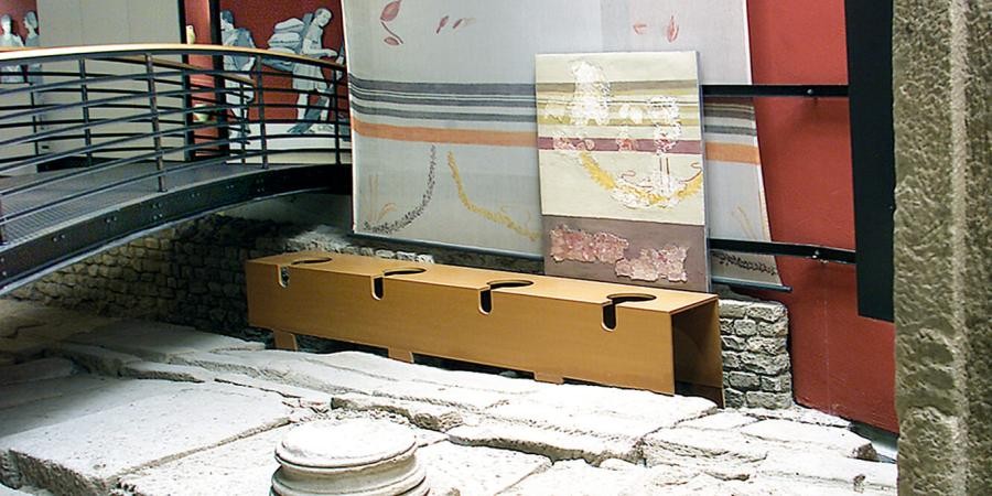 Ausschnitt der Latrinenanlage im Sumelocenna-Museum Rottenburg, mit Abflusskanal, darauf rekonstruierte Toilettensitzreihe und teilweise originale Reste der Wandbemalung, davor Steinboden und Säulenstumpf
