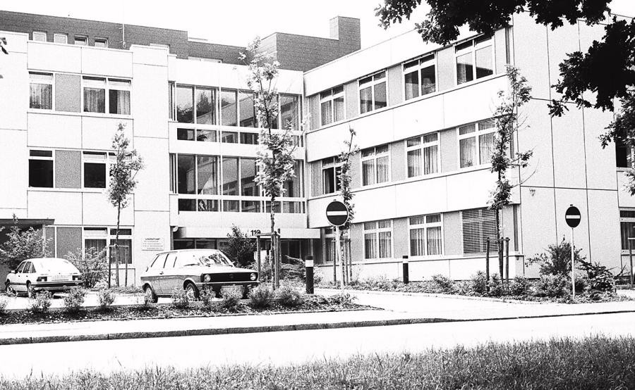 Dreistöckiges, flach gedecktes Gebäude, rechtwinklig angeordnet, davor ein Parkplatz und eine Straße