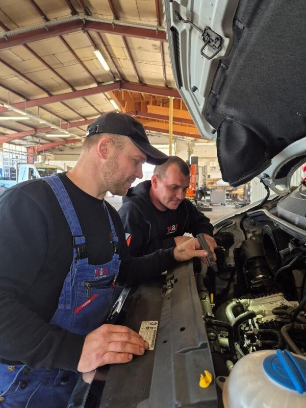 Zwei Personen in Arbeitskleidung schauen in einer Werkstatt in den Motorraum eines Fahrzeugs, einer leuchtet mit der Taschenlampe hinein,