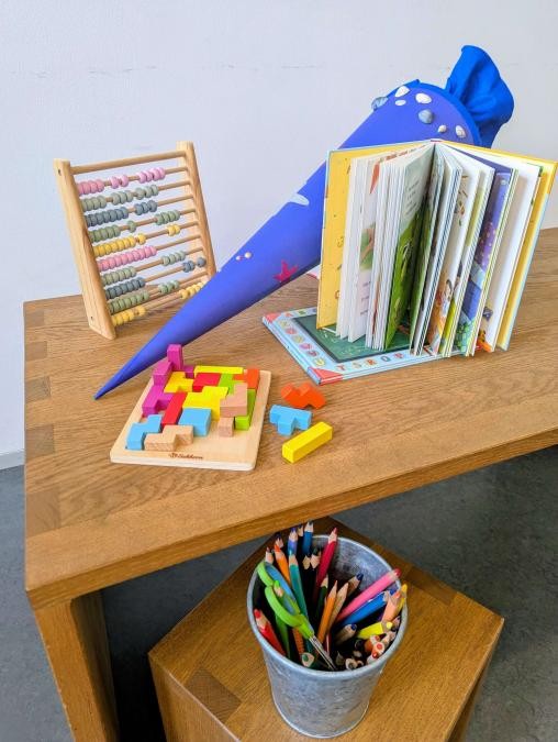 Ein Schreibtisch mit Hocker, darauf eine Schultüte, ein Buch, ein Rechenschieber, ein Holzpuzzle und ein Behälter mit Buntstiften