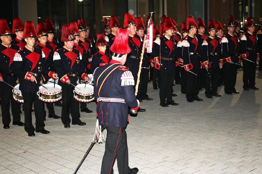 Mehrere Reihen von Männern stehen hintereinander, in Uniform mit auffälligem Helmbusch und mit Musikinstrumenten, davor schreitet ein uniformierter Mann mit Fahne die Reihenab