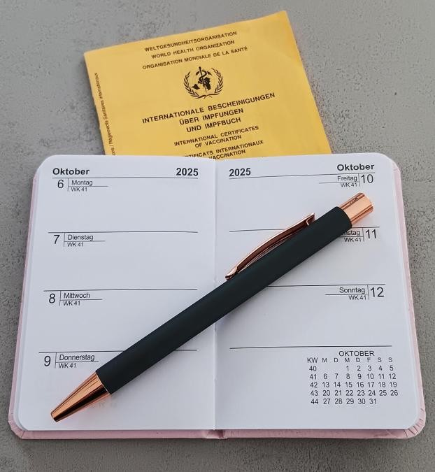 Aufgeschlagener Kalender darauf ein Kuli, unter dem Kalender liegt ein Impfpass