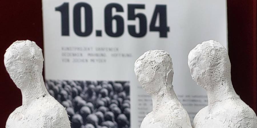 Im Vordergrund sind drei weiße Gipsfiguren bis zur Brust zu sehen. Im Hintergrund ist ist ein Banner, auf dem in fetter schwarzer Schrift die Zahl 10.654 zu sehen ist 