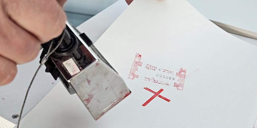 eine Hand hält einen Stempel, auf einem Blatt ein Stempel mit einem roten Kreuz 