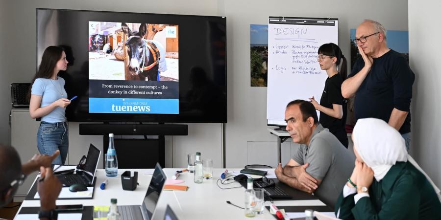 Mehrere Personen um einen Tisch, zwei jüngere Personen stehen an einem Bildschirm und einem Flipchart
