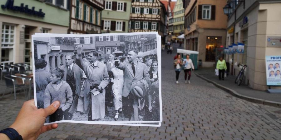 Blick in eine Straße der Tübinger Altstadt, davor eine Hand die Fotografien ins Bild hält mit schwarz-weiß-Aufnahmen einer Menschenmenge, im Vordergrund zeigen Männer den Hitlergruß