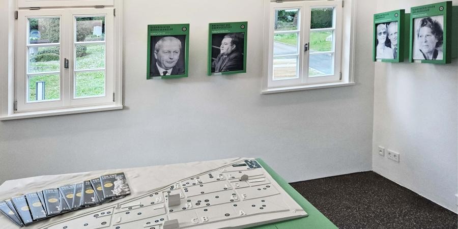 Ausstellung im ehemaligen Friedhofswärterhaus: Innenraum mit Blick auf den Stadtfriedhof, an den Wänden Fotografien von Personen, auf dem Tisch ein Lagemodell des Stadtfriedhofs und Faltblätter