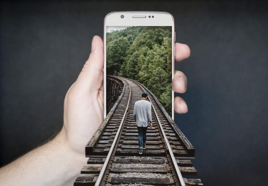 Eine Hand hält ein Handy, in das Handy hinein führen Gleise auf denen eine Person auf eine Brücke immer weiter gehtr
