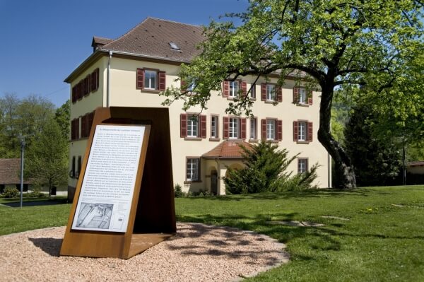 Dreistöckiges Gebäude mit Walmdach in einem parkähnlichen Garten, davor auf einem Eisenträger eine Gedenktafel