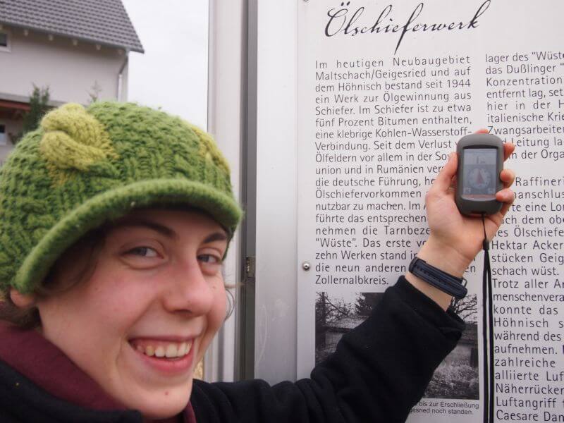 Geocache Dußlingen Junge Frau hält ein Geocache-Gerät in der Hand, sie steht vor einer Infotafel mit der Überschrift "Ölschieferwerk"